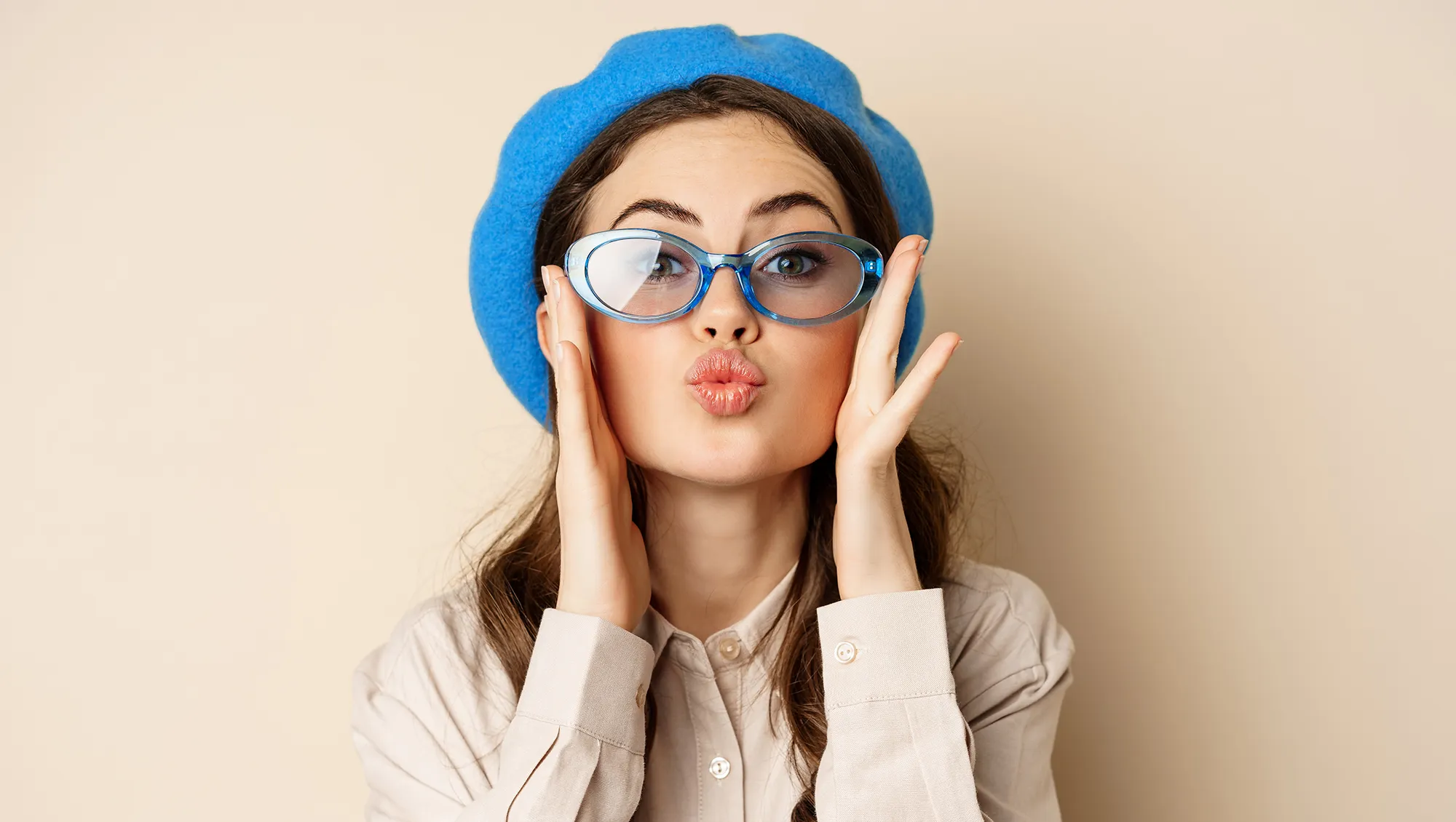 Beautiful stylish woman in sunglasses, blowing air kiss on hands near lips, gazing softly and flirty at camera, posing against beige background.