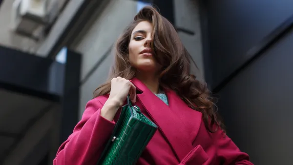 Fabulous portrait of the young stylish brunette. Elegant young woman with long wavy dark hair looks down thoughtfully. She wears stylish crimson coat and green leather bag.