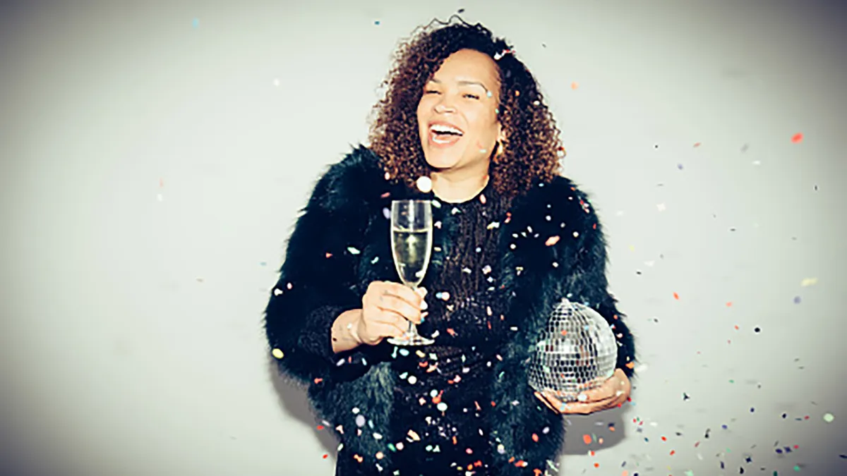 A smiling woman is holding a glass of champagne and a disco ball, wearing a black party dress amid falling confetti against a spot lit white background
