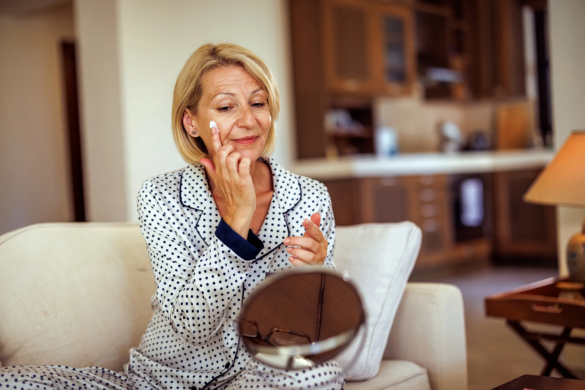 Smiling mature woman applying face cream under her eye while doing skin care routine at home