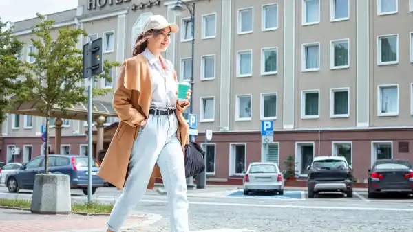 Afternoon stroll: Confident young lady with casual clothing style strolls along sunlit paved sidewalk in downtown, taking sips from coffee cup, crossing road and embracing urban charm around and nice weather