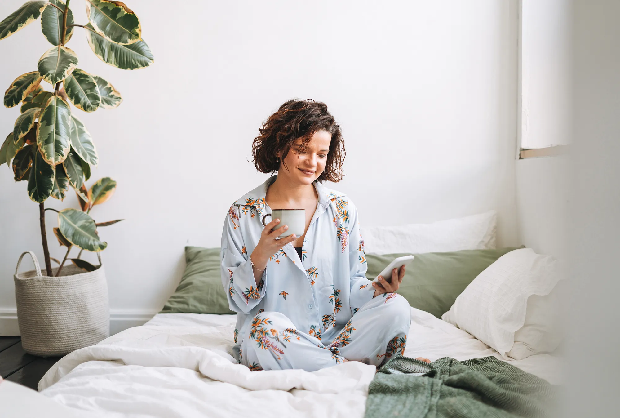 Young brunette woman with curly hair in pajamas using mobile on bed at home