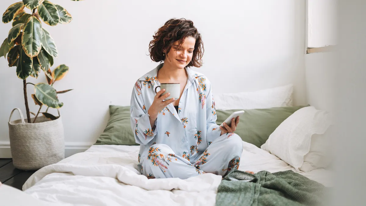 Young brunette woman with curly hair in pajamas using mobile on bed at home