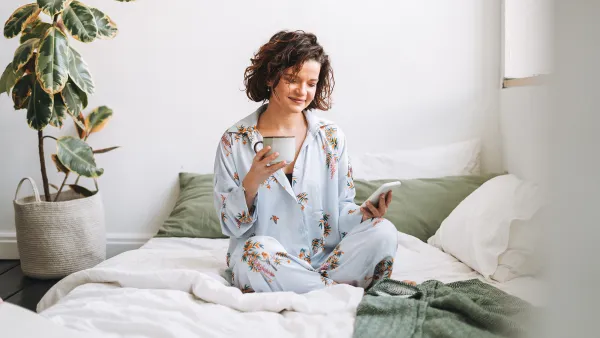 Young brunette woman with curly hair in pajamas using mobile on bed at home
