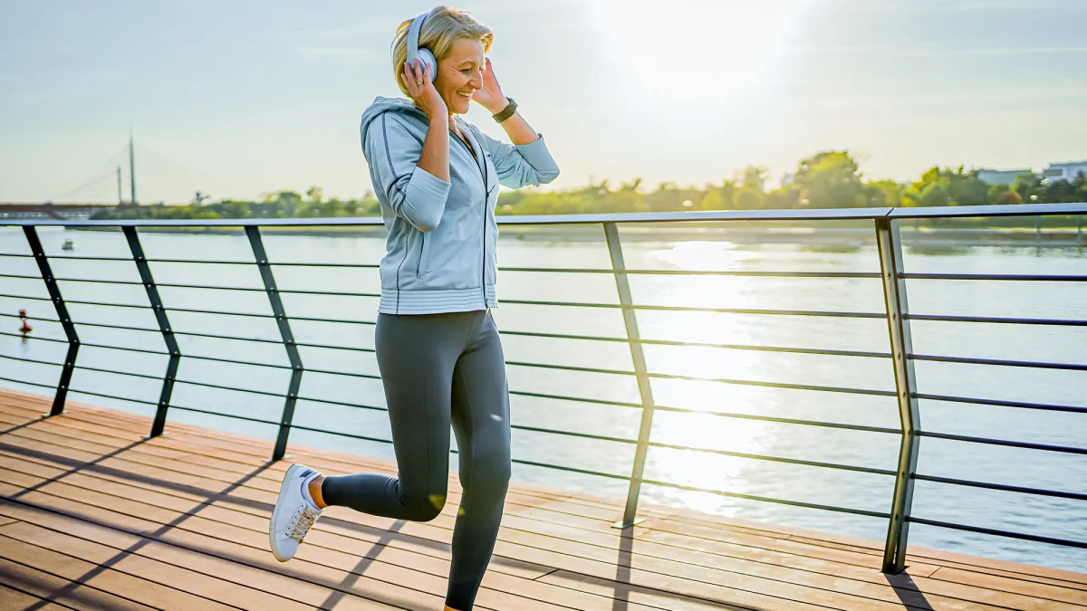 Cheerful mature woman with headphones jogging on the riverside