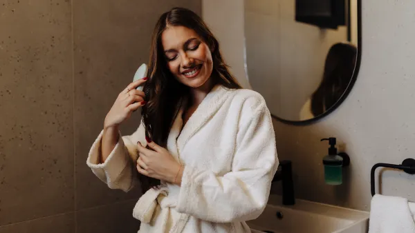 Woman Combing Her Beautiful Hair With Brush While Standing Near Mirror In Bathroom