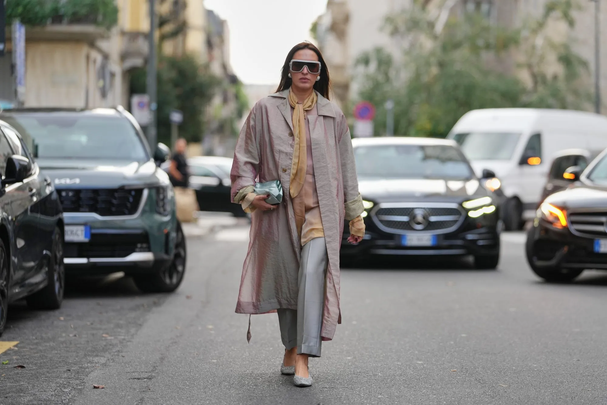 MILAN, ITALY - SEPTEMBER 23: A guest wears long straight dark brown hair parted in the center, oversized white rectangular sunglasses with dark lenses, large silver circular earrings, a small metallic pale aqua leather pouch clutch held in the hand, a long iridescent pale pink trench coat with button front and long belt ties and rolled cuffs showing a pale green lining, a golden yellow silk scarf tied loosely at the neck over a beige silk blouse, cropped light grey tailored trousers, grey textured pointed-toe flats, outside Alberta Ferretti, during Milan Fashion Week - WoWomenswear Spring/Summer 2026, on September 23, 2025 in Milan, Italy (Photo by Edward Berthelot/Getty Images)