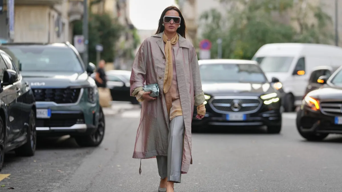 MILAN, ITALY - SEPTEMBER 23: A guest wears long straight dark brown hair parted in the center, oversized white rectangular sunglasses with dark lenses, large silver circular earrings, a small metallic pale aqua leather pouch clutch held in the hand, a long iridescent pale pink trench coat with button front and long belt ties and rolled cuffs showing a pale green lining, a golden yellow silk scarf tied loosely at the neck over a beige silk blouse, cropped light grey tailored trousers, grey textured pointed-toe flats, outside Alberta Ferretti, during Milan Fashion Week - WoWomenswear Spring/Summer 2026, on September 23, 2025 in Milan, Italy (Photo by Edward Berthelot/Getty Images)