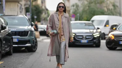 MILAN, ITALY - SEPTEMBER 23: A guest wears long straight dark brown hair parted in the center, oversized white rectangular sunglasses with dark lenses, large silver circular earrings, a small metallic pale aqua leather pouch clutch held in the hand, a long iridescent pale pink trench coat with button front and long belt ties and rolled cuffs showing a pale green lining, a golden yellow silk scarf tied loosely at the neck over a beige silk blouse, cropped light grey tailored trousers, grey textured pointed-toe flats, outside Alberta Ferretti, during Milan Fashion Week - WoWomenswear Spring/Summer 2026, on September 23, 2025 in Milan, Italy (Photo by Edward Berthelot/Getty Images)