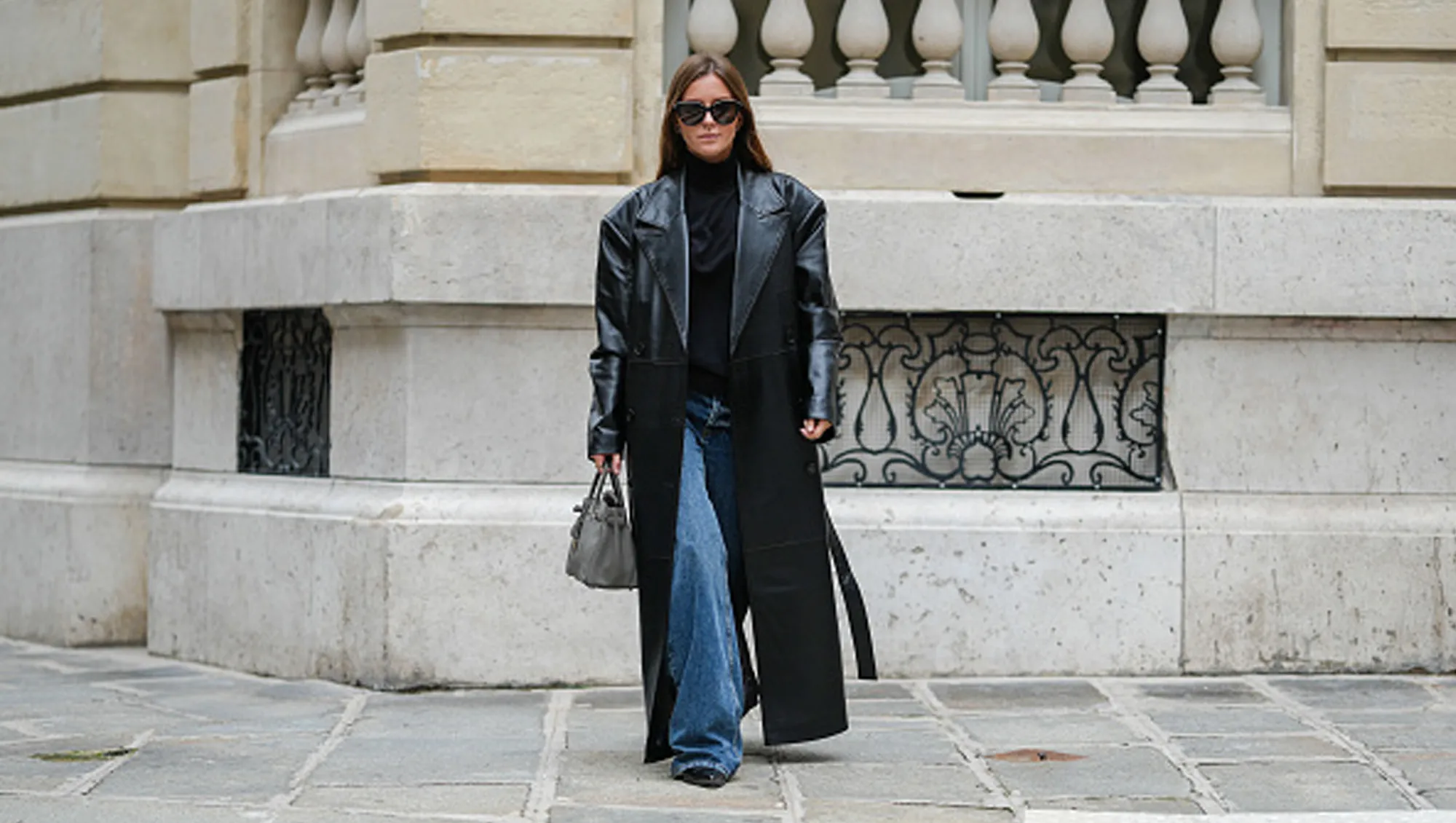 Diane Batoukina wears black Saint Laurent sunglasses, a black Tods turtleneck, a black faux leather long The Frankie Shop trench coat, a grey leather Birkin Hermes bag, blue denim jeans / pants, black leather Bershka shoes, during a street style fashion photo session, on October 18, 2024 in Paris, France.