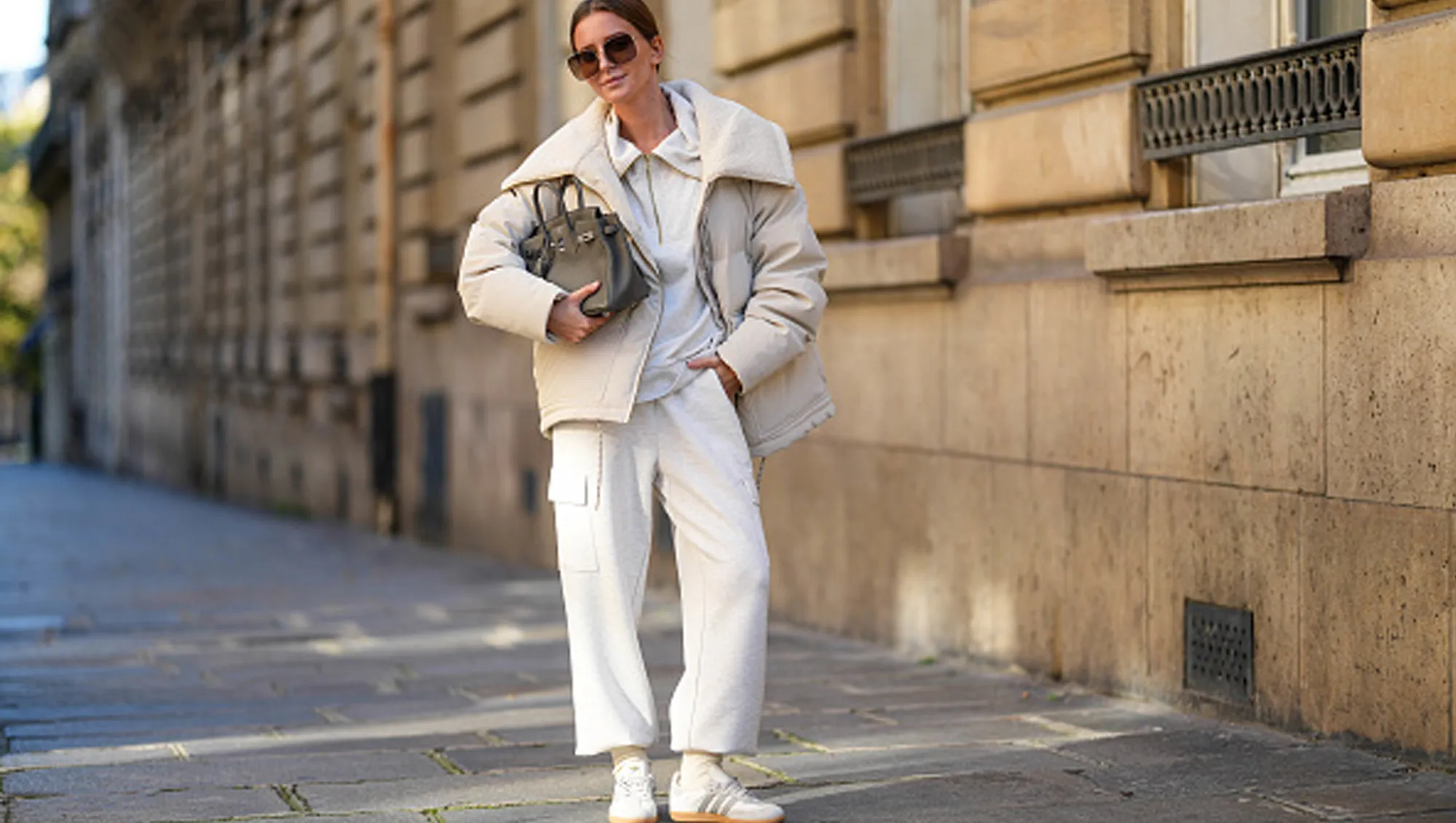 Diane Batoukina wears black Dior sunglasses, a white Varley puffer jacket, a white Adidas sweatshirt, white Varley pants, a grey leather Birkin Hermes bag, white socks, white Samba Adidas sneakers shoes, during a street style fashion photo session, on October 23, 2024 in Paris, France.