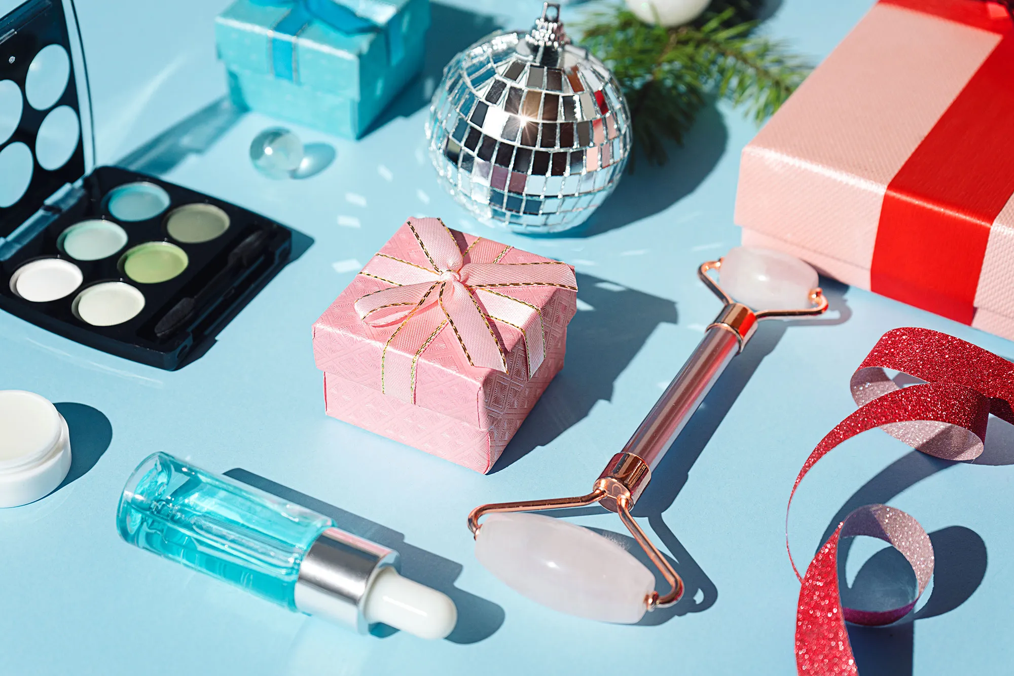 Pink Christmas gift box, rose quartz Jade face roller, cream, face serum, silver Disco balls, fir tree branch, trendy 2022 color eyeshadow, on pastel blue color background. Shiny glittering template for beauty product or cocktail party invitation. Holiday party, Merry Christmas, Happy New Year, Black Friday, Valentine's day and Birthday concept. Minimalistic background with copy space. Close-up. Flat lay.