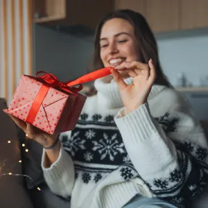 Beautiful woman holding a wrapped gift in her hand, opening it and smiling. A Christmas surprise or birthday gift from a friend delivered by mail.