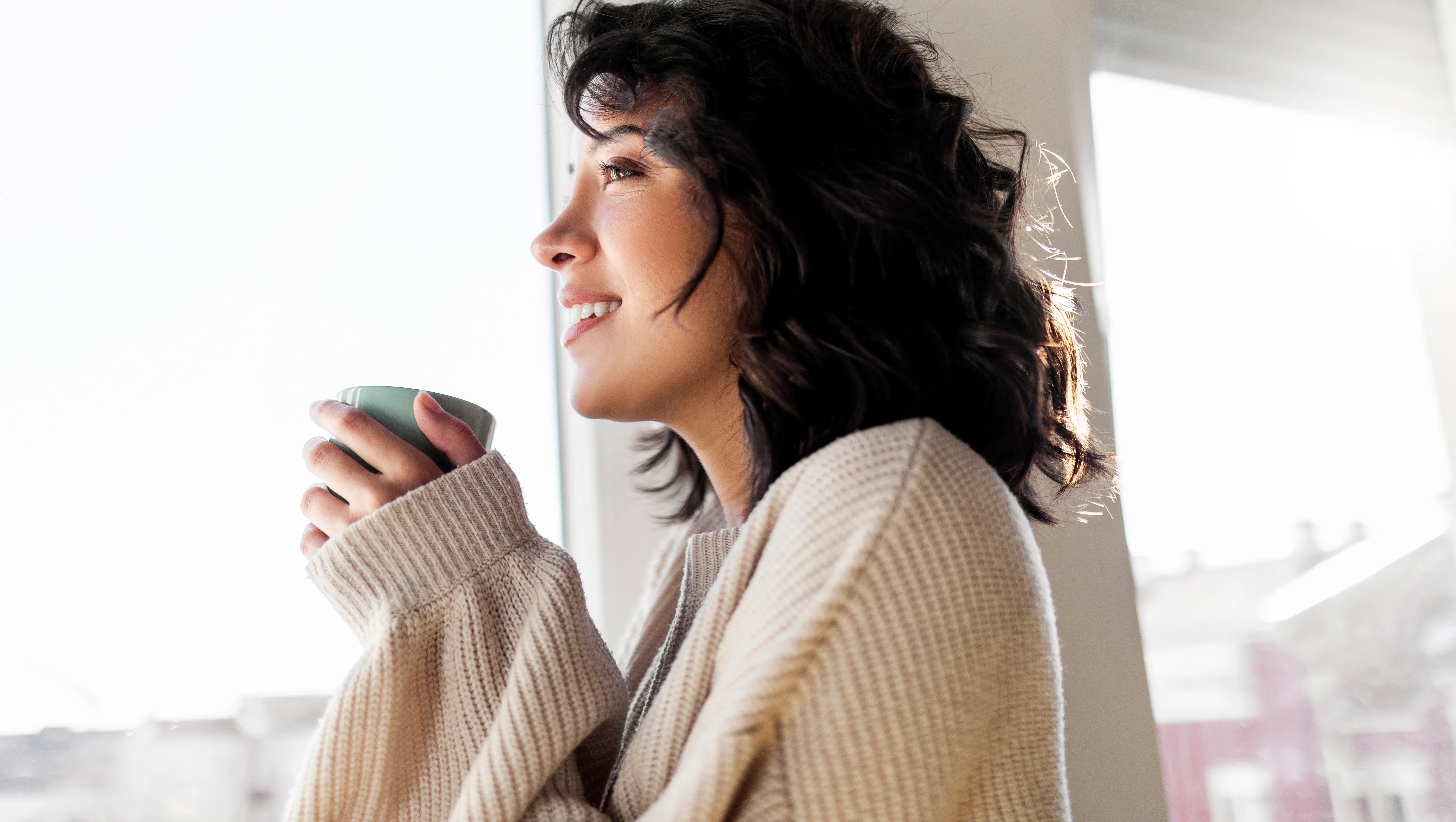 Beautiful young woman drinking cup of coffee while looking through window at home
