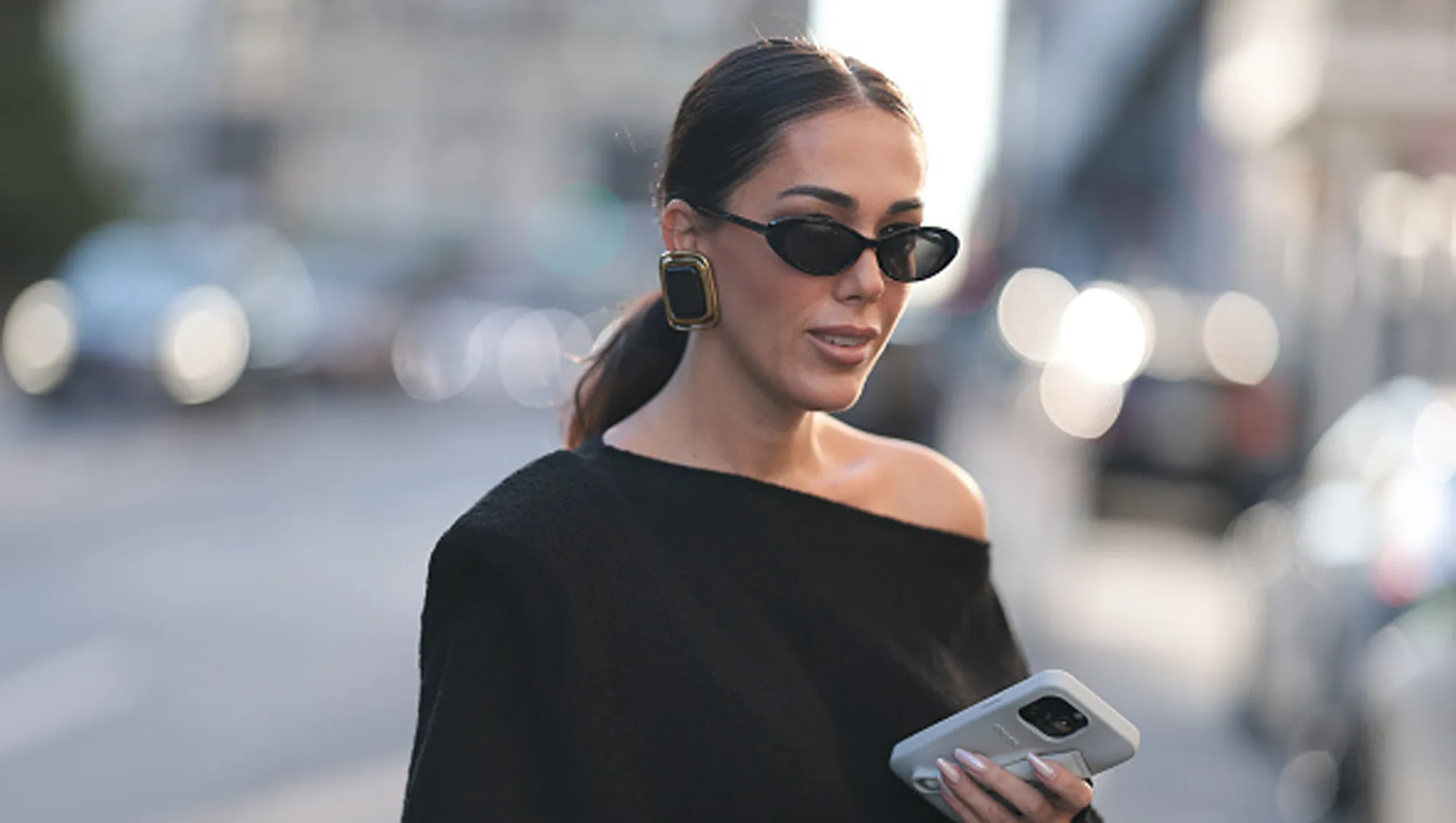 Olga Sokolenko seen wearing black sunglasses, Saint Laurent large gold / black square earrings, Say No More black off-shoulder asymmetrical oversized cotton sweater and Rhode grey silicone lip phone case, during the New York Fashion Week, on September 09, 2024 in New York City.
