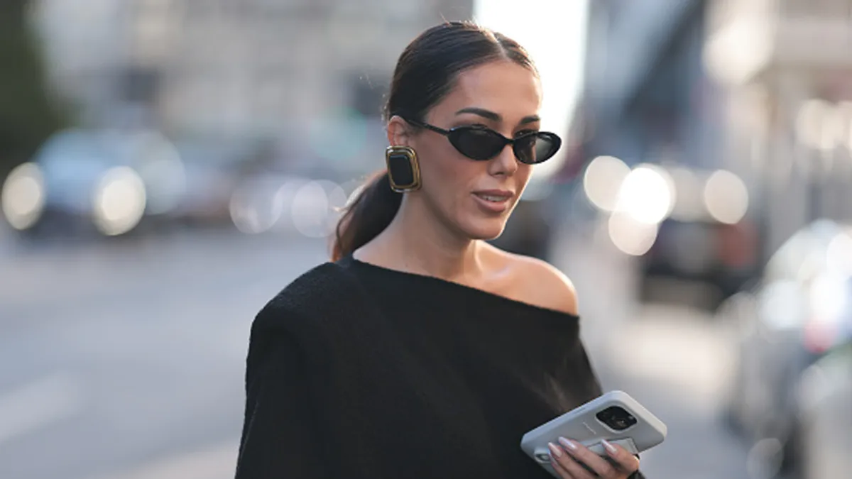 Olga Sokolenko seen wearing black sunglasses, Saint Laurent large gold / black square earrings, Say No More black off-shoulder asymmetrical oversized cotton sweater and Rhode grey silicone lip phone case, during the New York Fashion Week, on September 09, 2024 in New York City.