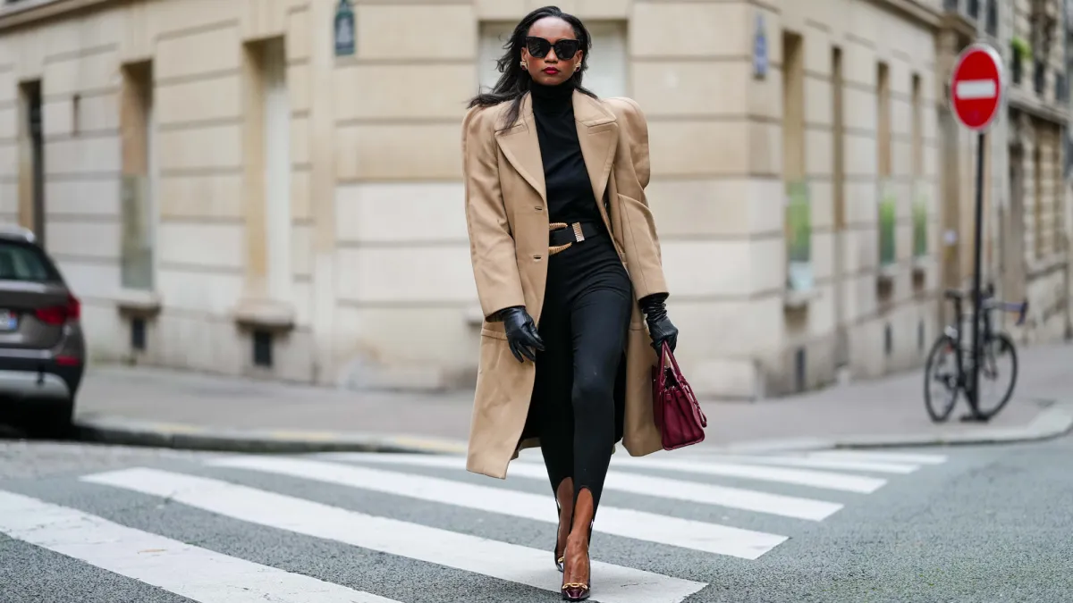 Emilie Joseph wears a wool camel tailored long coat from Givenchy Paris, wool black turtleneck from COS, high-waisted black stirrup pants / leggings from Mango, black leather belt wide Wide golden buckle, vintage black leather gloves, burgundy / cherry / oxblood pointed-toe heels featuring a subtle gold accent / horsebits, rich burgundy handbag from Saint Laurent Paris, black squared sunglasses from Gentle Monster, vintage style earrings with cherry hued stone, during a street style fashion photo session, on October 18, 2024 in Paris, France.