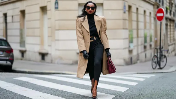 Emilie Joseph wears a wool camel tailored long coat from Givenchy Paris, wool black turtleneck from COS, high-waisted black stirrup pants / leggings from Mango, black leather belt wide Wide golden buckle, vintage black leather gloves, burgundy / cherry / oxblood pointed-toe heels featuring a subtle gold accent / horsebits, rich burgundy handbag from Saint Laurent Paris, black squared sunglasses from Gentle Monster, vintage style earrings with cherry hued stone, during a street style fashion photo session, on October 18, 2024 in Paris, France.