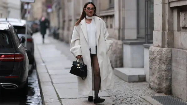 Anna Winter is seen wearing sunglasses with rimless oval grey lenses, a silver metal nose bridge, and transparent temples with silver logo detail from Chanel; chunky silver hoop earrings; a white slim wool scarf; an oversized ankle-length white coat with notch lapels; a white wool pullover with a V-neck underneath; grey shorts from The Frankie Shop; a black leather “Kelly” bag from Hermès with a black leather horse-shaped “Rodeo“ charm and gold cadena; a yellow gold “Love” ring, paired with a yellow gold “Juste un Clou” ring, both from Cartier; transparent black tights from Falke; and furry black ballerinas with a single strap from Scholl on November 29, 2024 in Berlin, Germany