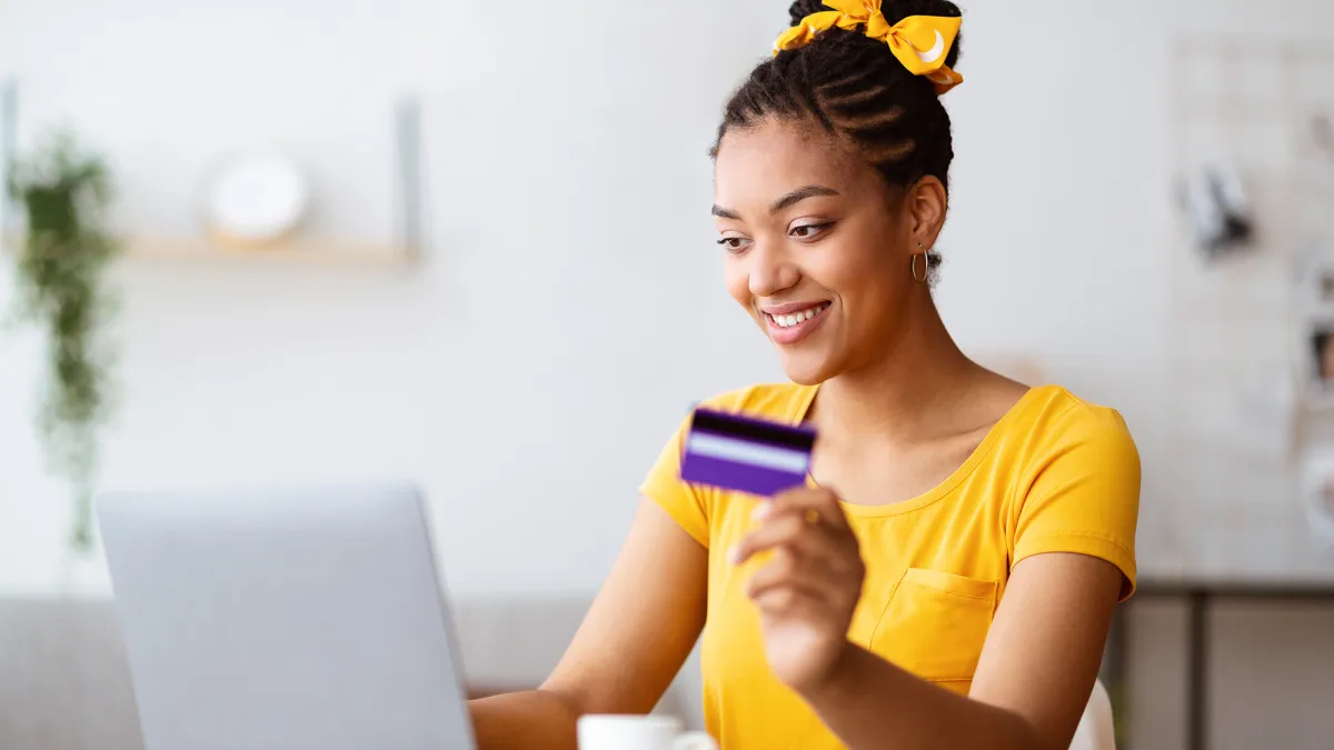 Online Shopping Concept. Portrait of smiling african american woman holding credit card in hand and using laptop, sitting at desk, typing on keyboard, blurred background, selective focus. Ecommerce