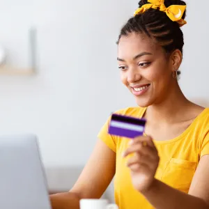 Online Shopping Concept. Portrait of smiling african american woman holding credit card in hand and using laptop, sitting at desk, typing on keyboard, blurred background, selective focus. Ecommerce