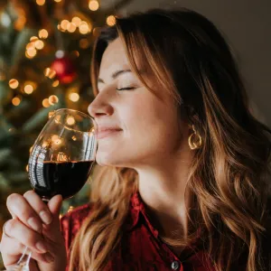 Woman smelling glass of wine beside decorated Christmas tree