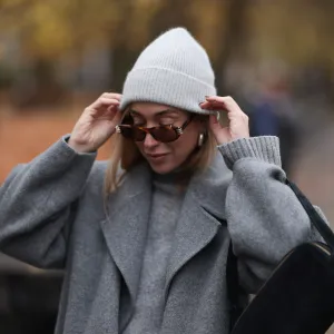 BERLIN, GERMANY - NOVEMBER 14: Sonia Lyson is seen wearing a light grey knit beanie from Le Bonnet; chunky silver hoop earrings; oval sunglasses with Havana frame and silver details from Givenchy; a calf-length grey wool coat with notch lapels from & Other Stories; a light grey knit sweater with ribbed turtleneck and cuffs from Sézane; and a rectangular black suede handbag from Elleme on November 14, 2024 in Berlin, Germany. (Photo by Jeremy Moeller/Getty Images)
