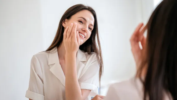 Happy beautiful millennial female apply nourishing under eye cream against wrinkles. Face lifting and anti aging cosmetic products. Smiling young woman use moisturizing balm, standing in bathroom