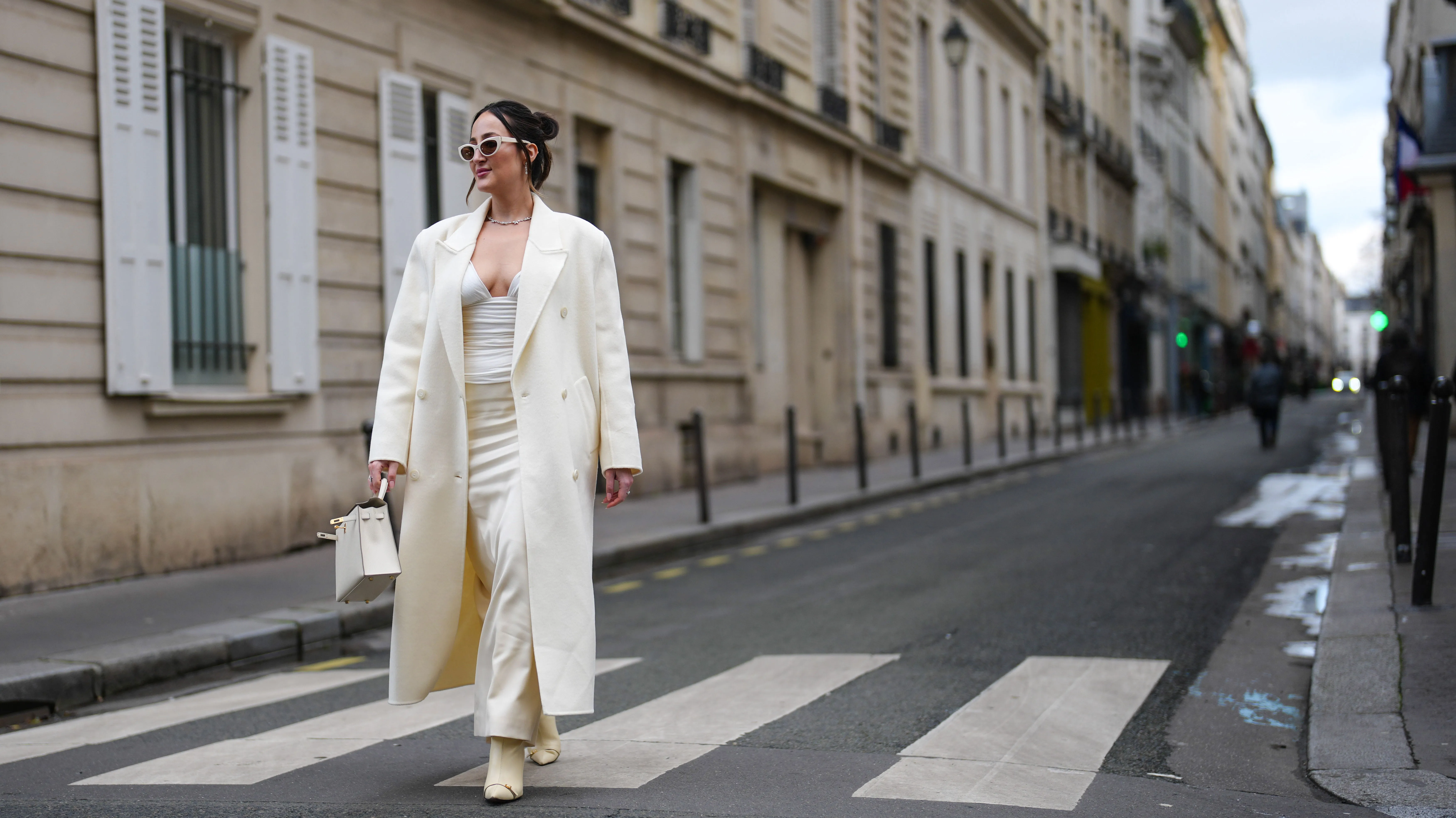 A guest wears cream sunglasses, silver necklace, white oversized double breasted coat, white loose body suit, cream pointed toe heeled boots, white Hermes Kelly leather bag, outside Miss Sohee, during the Haute Couture Spring/Summer 2025 on January 30, 2025 in Paris, France. (Photo by Edward Berthelot/Getty Images)