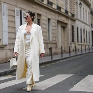 A guest wears cream sunglasses, silver necklace, white oversized double breasted coat, white loose body suit, cream pointed toe heeled boots, white Hermes Kelly leather bag, outside Miss Sohee, during the Haute Couture Spring/Summer 2025 on January 30, 2025 in Paris, France. (Photo by Edward Berthelot/Getty Images)