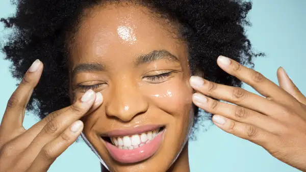 Close-up of woman applying glossy face make-up moisturizer, smiling, with eyes closed with turquoise background