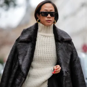 PARIS, FRANCE - JANUARY 21: A guest wears sunglasses, earrings, a white ribbed wool long pullover worn as a dress, a black fluffy long coat, outside Sacai, during the Menswear Fall/Winter 2024/2025 as part of Paris Fashion Week on January 21, 2024 in Paris, France. (Photo by Edward Berthelot/Getty Images)