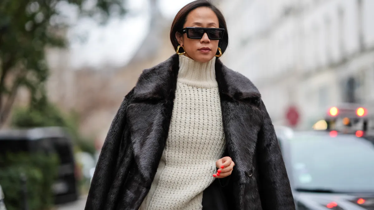 PARIS, FRANCE - JANUARY 21: A guest wears sunglasses, earrings, a white ribbed wool long pullover worn as a dress, a black fluffy long coat, outside Sacai, during the Menswear Fall/Winter 2024/2025 as part of Paris Fashion Week on January 21, 2024 in Paris, France. (Photo by Edward Berthelot/Getty Images)