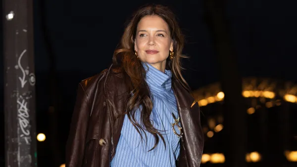 PARIS, FRANCE - JANUARY 26: Katie Holmes wears gold Patou earrings, white light blue striped long sleeve Patou shirt, gold Patou brooch, shiny dark brown double breasted Patou leather trench coat, dark brown straight Patou trouser pants, shiny dark blue Patou leather bag, black white polka dot heels, outside Patou, during the Paris Fashion week Men’s Fall/Winter 2025-2026 on January 26, 2025 in Paris, France. (Photo by Edward Berthelot/Getty Images)