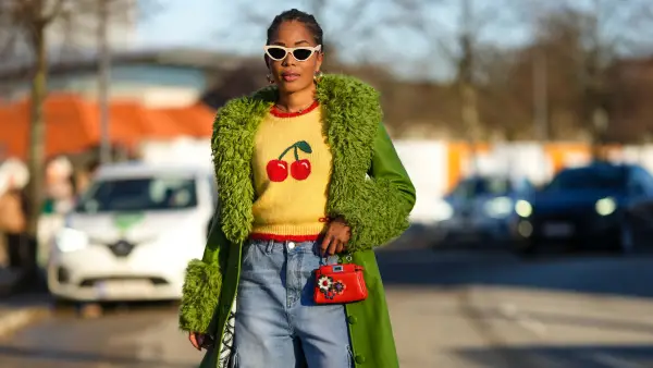 COPENHAGEN, DENMARK - FEBRUARY 02: Ellie Delphine wears white latte sunglasses, gold earrings, a yellow wool with embroi dered red cherry pattern pullover, a green shiny leather with green oversized sheep fur borders long coat, a red shiny leather micro handbag, blue faded denim large puffy pants, yellow shiny satin with black rhinestones buckle on the toe-cap pumps heels shoes , outside Munthe, during the Copenhagen Fashion Week Autumn/Winter 2023 on February 02, 2023 in Copenhagen, Denmark. (Photo by Edward Berthelot/Getty Images)