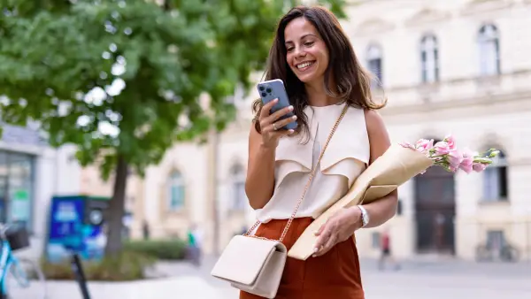 Woman smiling outdoors holding flowers and using phone
