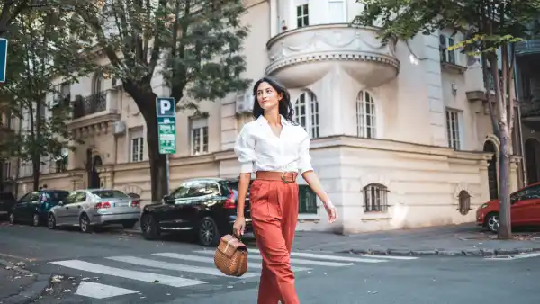 A stylish woman walks confidently down a picturesque street in a charming urban neighborhood, exuding elegance and freedom. She carries a woven handbag and is surrounded by classic architecture and parked cars.