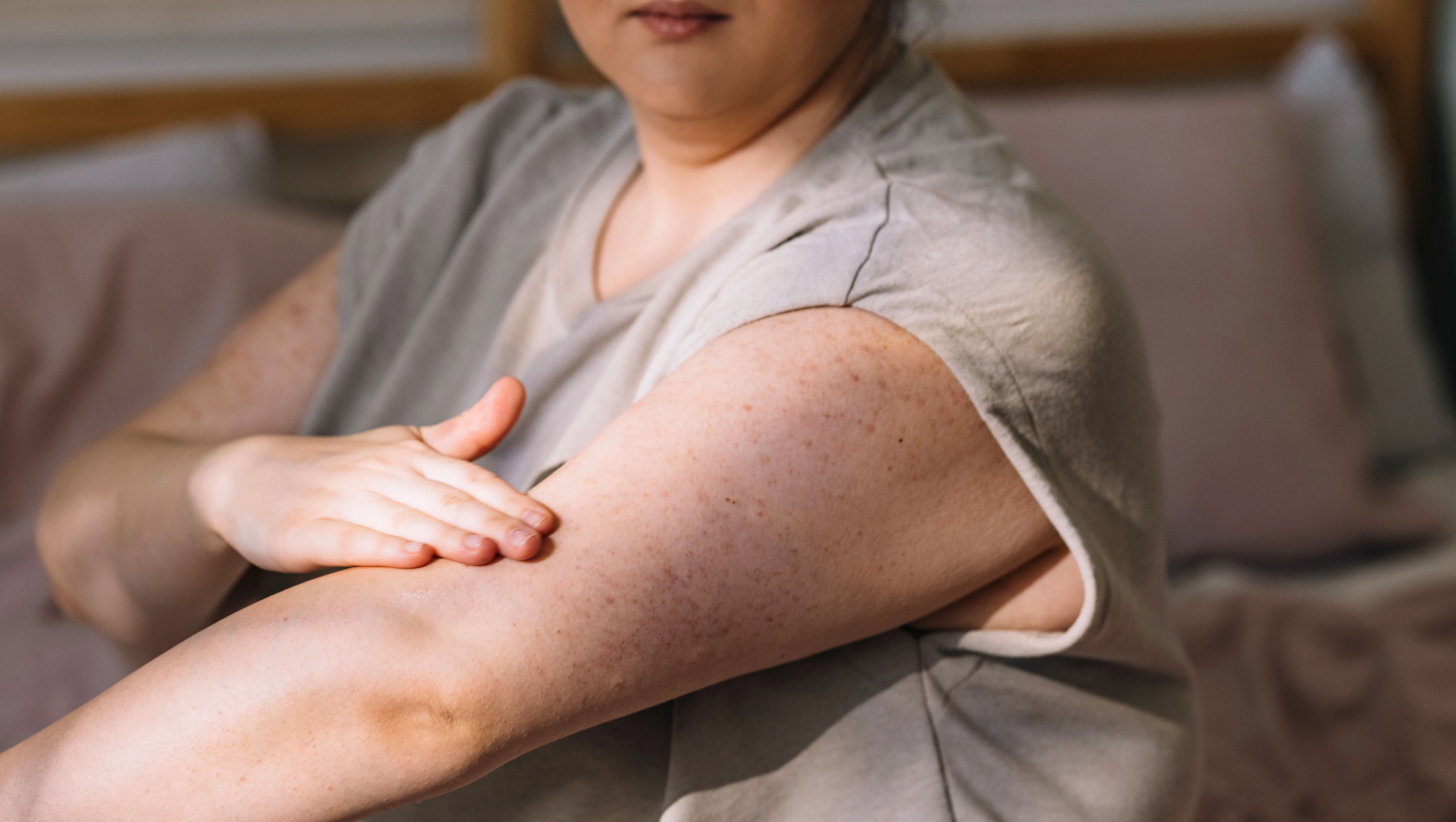 A cropped image of a plus size young woman sitting on her bed in the morning and applying body lotion on her arms