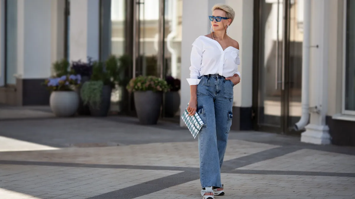 Rejuvenated Trendy mature woman with short hair stands outdoors in city street wears youth clothing white shirt, jeans cargo pants, clutch. Urban style fashion, age and timeless beauty.