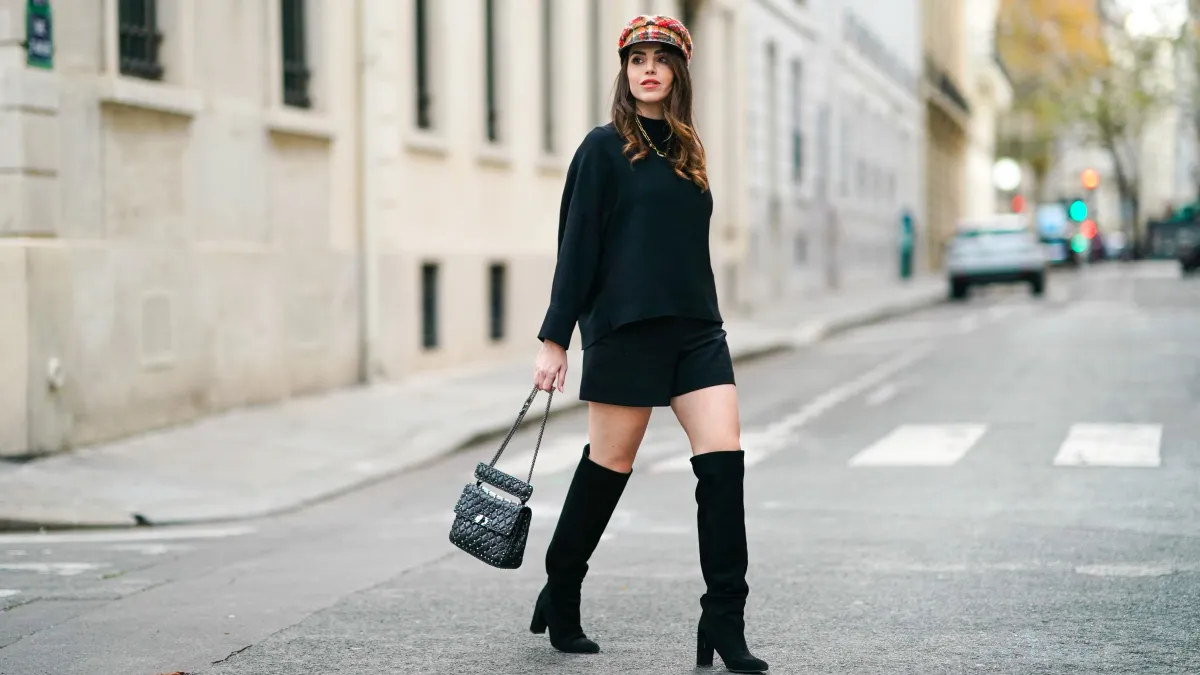 PARIS, FRANCE - NOVEMBER 14: Sara Carnicella wears a red checked beret hat from Anthony Peto, a black oversized pullover from Natan, a black leather studded Valentino Rockstud bag, Zara black shorts, black pointy knee-high boots from Jonak, a golden necklace from APM Monaco, on November 14, 2020 in Paris, France.