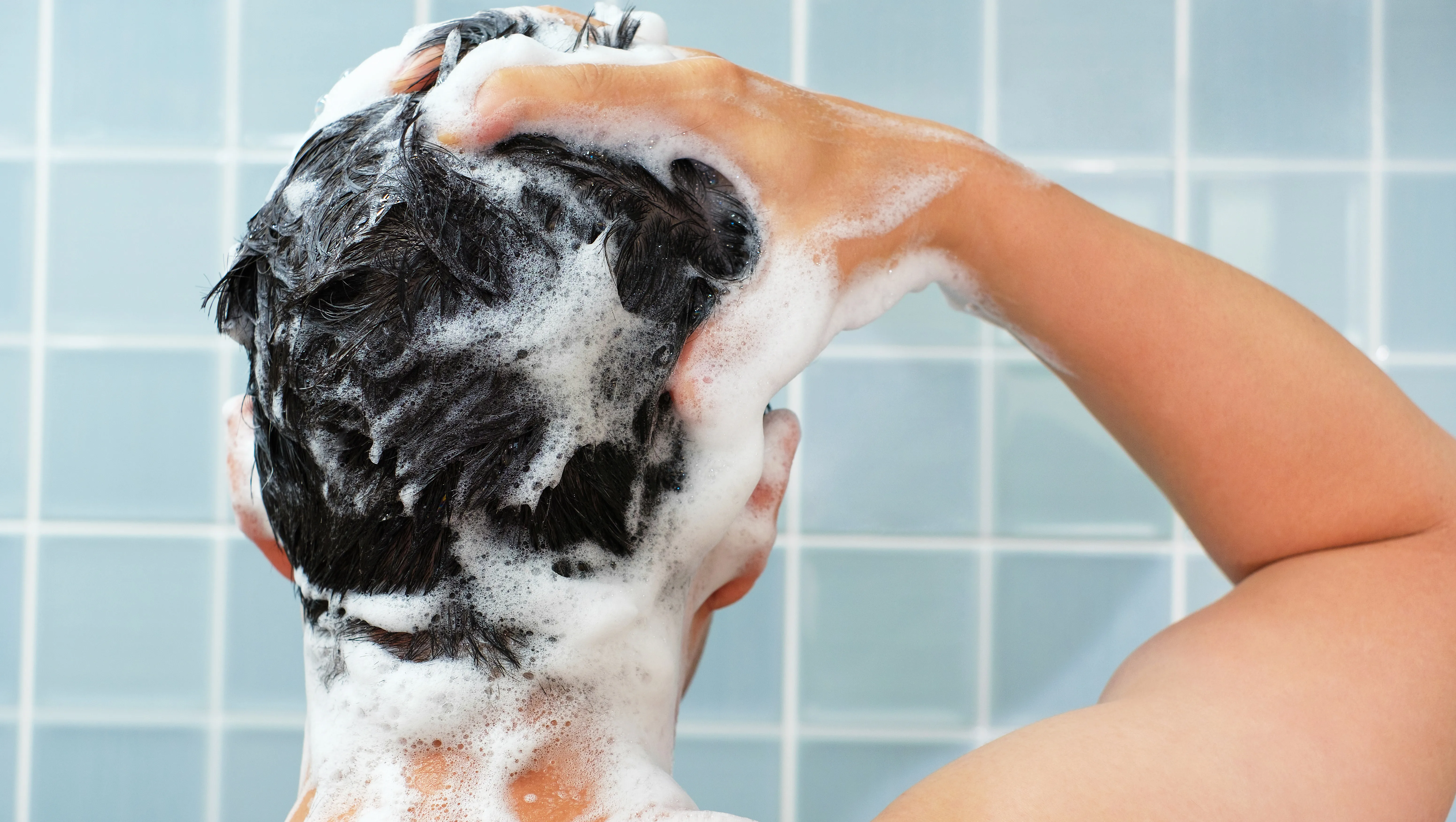 Male hands wash their hair with shampoo and foam on a blue background, rear view.
