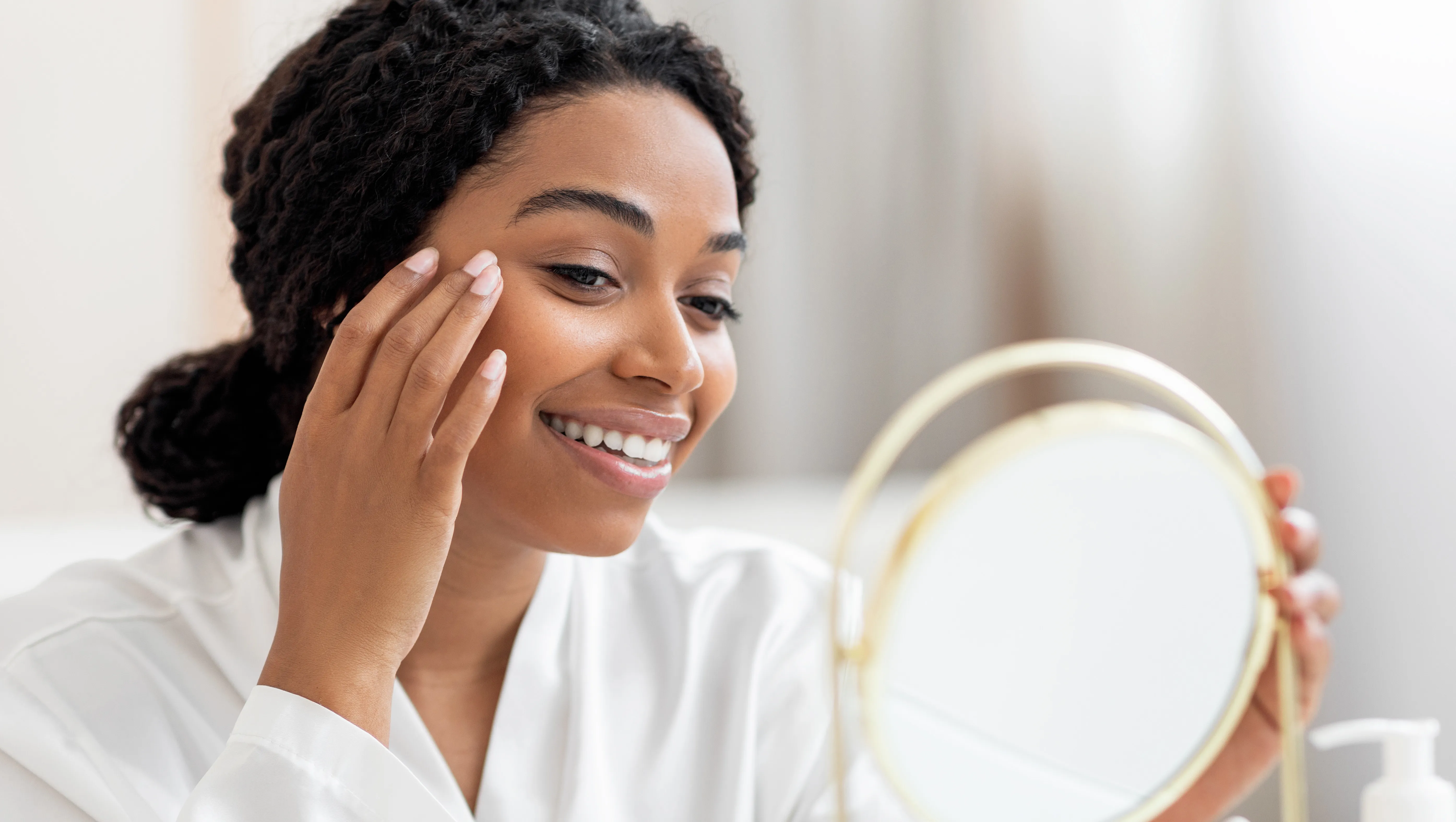 Beautiful Young Black Lady Looking In Mirror And Applying Under Eye Cream, Happy Smiling African American Woman In White Silk Robe Making Beauty Treatments At Replica Luxury Handbag, Enjoying Self-Care, Closeup