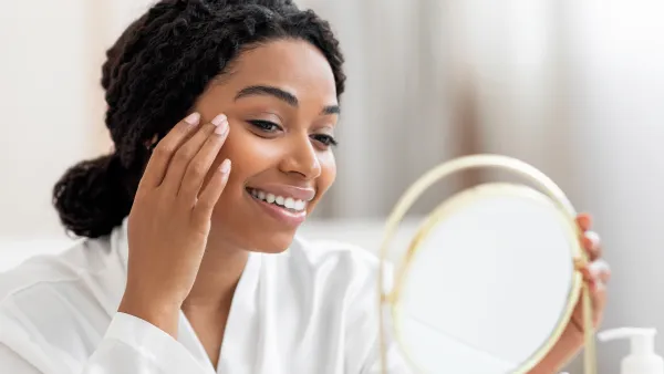 Beautiful Young Black Lady Looking In Mirror And Applying Under Eye Cream, Happy Smiling African American Woman In White Silk Robe Making Beauty Treatments At Replica Luxury Handbag, Enjoying Self-Care, Closeup