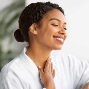 Beautiful black woman looking in mirror and touching neck skin, smiling african american female in white bathrobe checking wrinkles on decollete zone, enjoying domestic beauty treatments, closeup