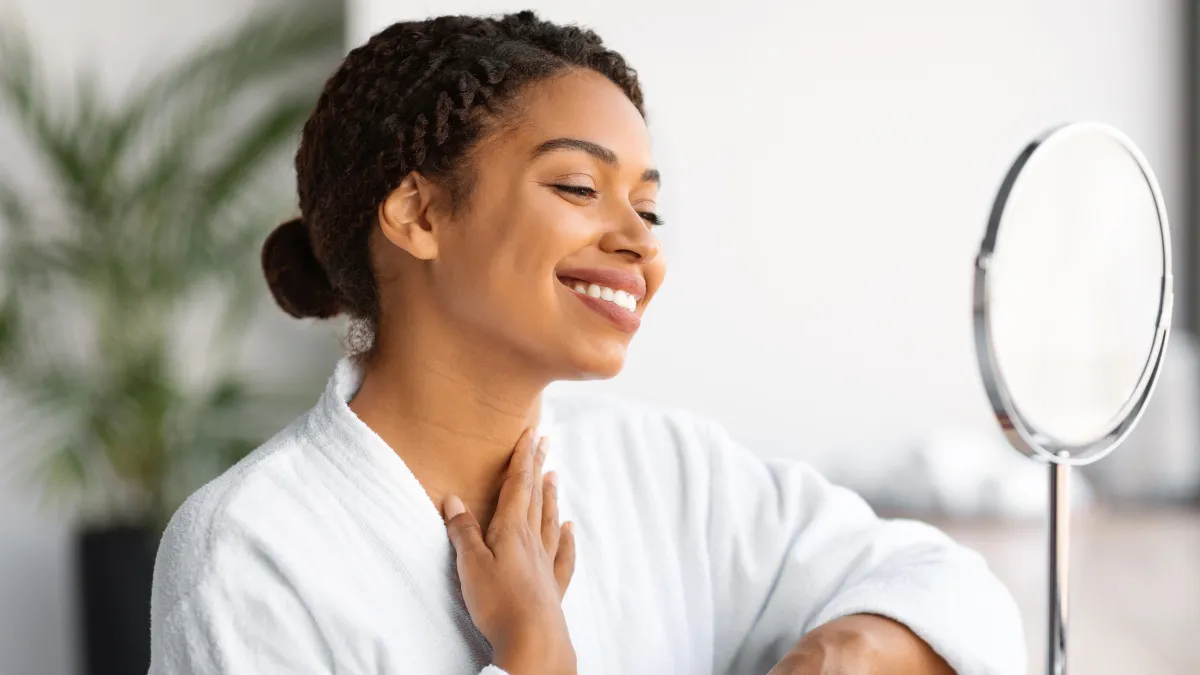 Beautiful black woman looking in mirror and touching neck skin, smiling african american female in white bathrobe checking wrinkles on decollete zone, enjoying domestic beauty treatments, closeup