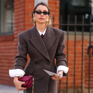 NEW YORK, NEW YORK - FEBRUARY 07: A guest wears black sunglasses, gold earrings, white buttoned up long sleeve shirt, burgundy bordeaux tie, dark brown double breasted oversized tailored blazer jacket, burgundy bordeaux leather clutch bag with gold chain strap, dark gray socks, shiny black gold detail loafer moccasin leather shoes, outside Theory, during the New York Fashion week Fall/Winter 2025 on February 7, 2025 in New York, New York. (Photo by Edward Berthelot/Getty Images)