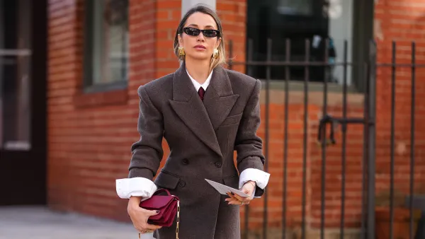 NEW YORK, NEW YORK - FEBRUARY 07: A guest wears black sunglasses, gold earrings, white buttoned up long sleeve shirt, burgundy bordeaux tie, dark brown double breasted oversized tailored blazer jacket, burgundy bordeaux leather clutch bag with gold chain strap, dark gray socks, shiny black gold detail loafer moccasin leather shoes, outside Theory, during the New York Fashion week Fall/Winter 2025 on February 7, 2025 in New York, New York. (Photo by Edward Berthelot/Getty Images)