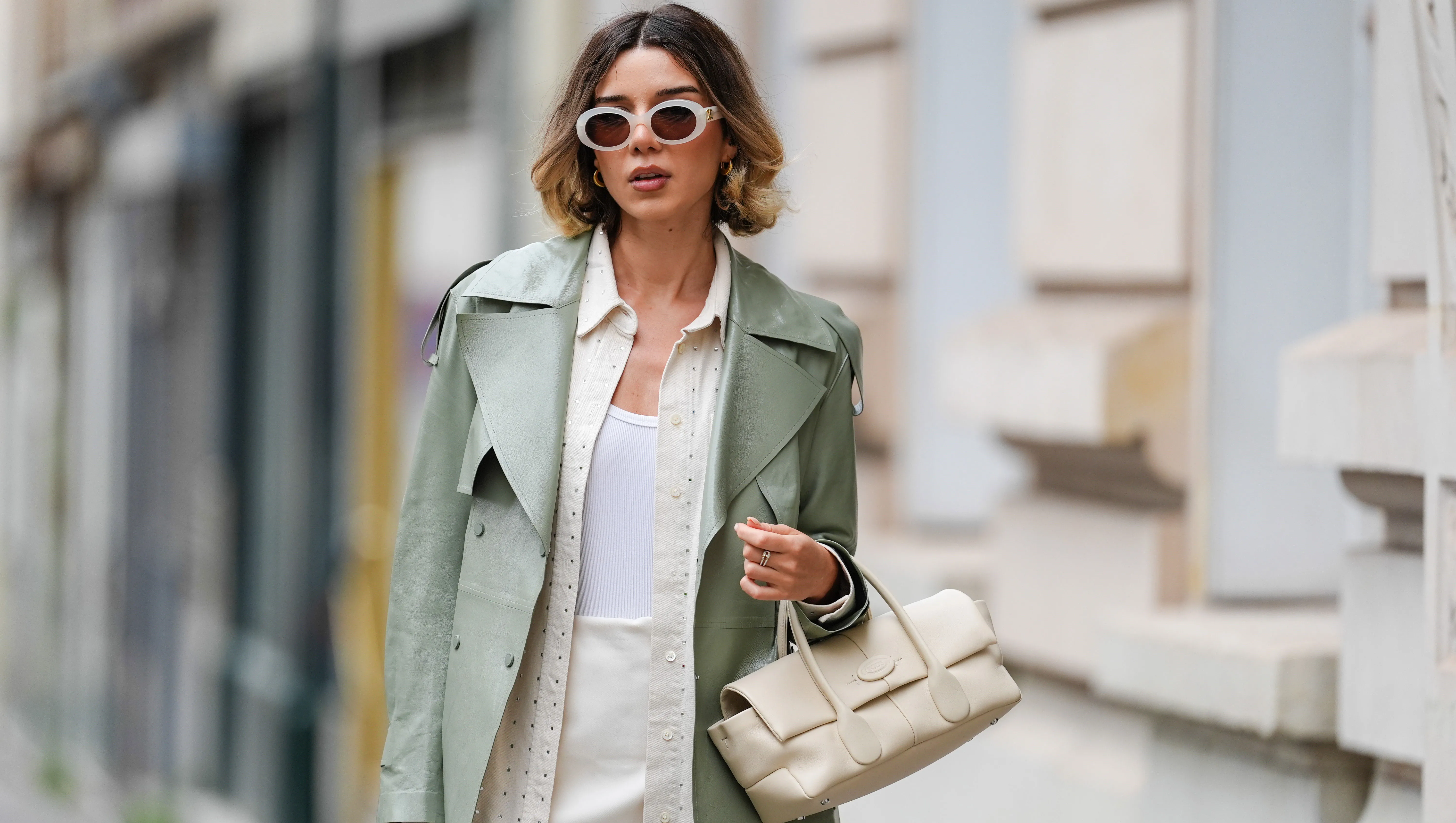 PARIS, FRANCE - APRIL 07: Katie Giorgadze wears sunglasses from Celine, golden earrings, a sage pastel green smooth leather trench coat from Mackage, a cream colored mini skirt, a white tank top from Uniqlo, a beige oversize rhinestone sparkly leather shirt from Frame, a cream color / white leather bag from Tod's, during a street style fashion photo session, on April 07, 2024 in Paris, France.