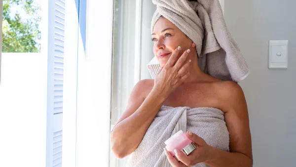 Smiling woman applying moisturizer on face standing by window at home