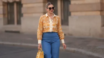 BERLIN, GERMANY - JUNE 07: Aline Kaplan is seen wearing large sunglasses with beige, brown and black pattern from Tibi; chunky golden hoop earrings; several golden necklaces; a creme-white blouse with floral orange pattern from Sézane; a blue high-waisted pair of trousers with two button borders on the hips from Sézane; golden rings and a yellow leather handbag from Saint Laurent on June 07, 2024 in Berlin, Germany.